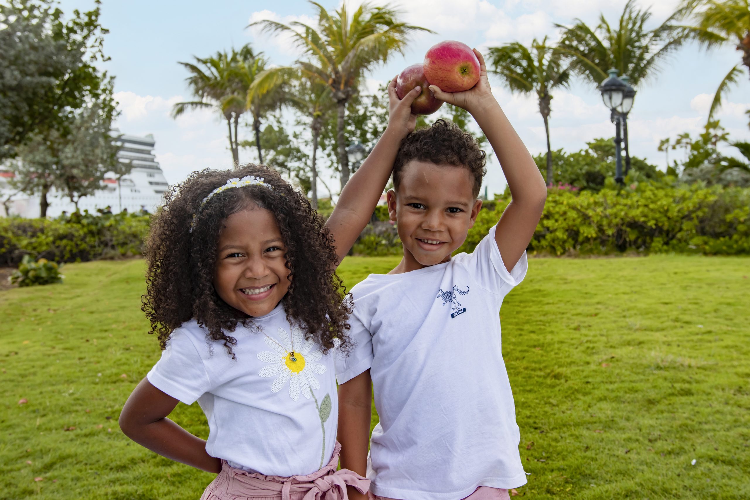 Two kids on a patch of grass are holding an apple above their head while smiling at the camera